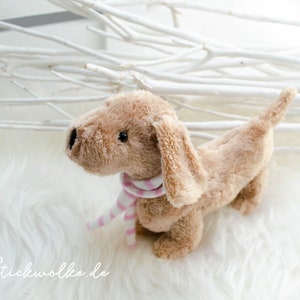 Puede incluir: Un juguete de peluche de dachshund marrón que lleva una bufanda de rayas rosa y blancas. El juguete está sentado sobre una superficie blanca y esponjosa.