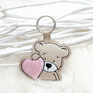 Può includere: Un portachiavi in ​​pelle color oro con un orsacchiotto marrone che tiene un cuore rosa. Il portachiavi ha un anello per chiavi argentato e il testo "STICKWOLKE.DE" è visibile in basso.