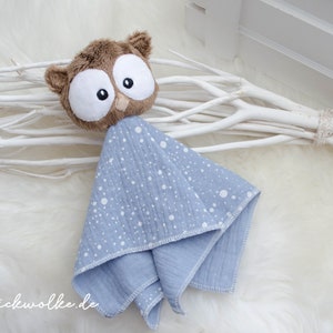 May include: A blue and white muslin baby blanket with a brown owl attached. The blanket has white polka dots on a light blue background. The owl has large brown eyes and a brown beak.
