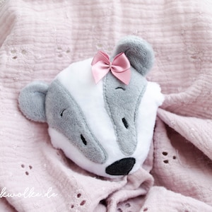 Könnte beinhalten: Ein Plüschtier in Form eines grauen und weißen Dachses mit einer rosa Schleife auf dem Kopf. Das Spielzeug liegt auf einer rosa Decke mit einem weißen Spitzenmuster.