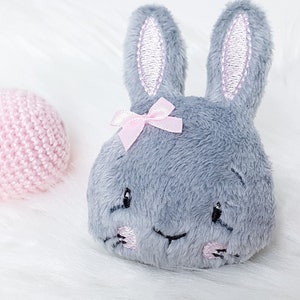May include: A gray plush bunny with pink ears and a pink bow. The bunny has embroidered eyes and a smile.