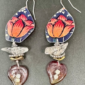 May include: A pair of dangle earrings featuring a floral design. The top part of the earrings is dark blue with a red flower. The bottom part of the earrings has a clear crystal, a gold bead, and a dark red heart-shaped charm.