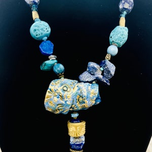May include: A blue and gold necklace with a large, carved pendant. The pendant is made of blue and gold ceramic and features a floral design. The necklace is made of various blue and gold beads, including lapis lazuli, turquoise, and gold.