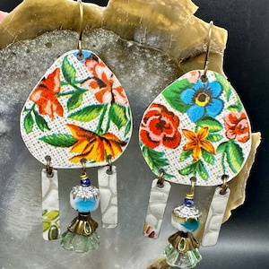 May include: A pair of floral patterned earrings with dangling charms. The earrings are made of metal and feature a white background with colourful flowers and green leaves. The charms are made of metal and feature a white background with a floral pattern. The earrings are a unique and stylish accessory.