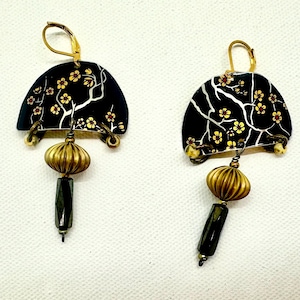 May include: A pair of handcrafted earrings. Each earring features a black, semi-circular top with a floral design in yellow and white. Below, a gold-colored, ribbed bead hangs, followed by a dark, rectangular bead. The earrings have gold-colored ear wires.