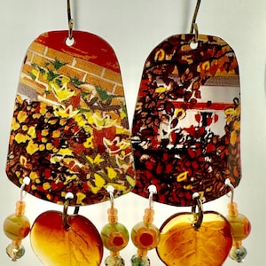 May include: A pair of dangle earrings with a red, orange, yellow and green floral pattern on the top portion. The bottom portion of the earrings feature a heart-shaped amber leaf with clear and green beads.