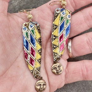 May include: A pair of colourful, geometric patterned earrings with gold accents. Each earring features a small, round charm with a face on it.