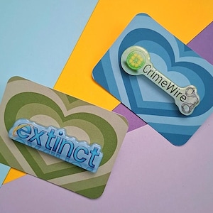 Könnte beinhalten: Zwei rechteckige Karten mit herzförmigen Designs und Anstecknadeln. Eine Anstecknadel sagt "extinct" in Blau, die andere sagt "CrimeWire" mit einem Limettendesign und Handschellen. Der Hintergrund ist eine Mischung aus Blau, Gelb, Grün und Lila.