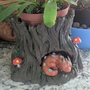 May include: A brown tree stump planter featuring a textured bark surface, two small red mushrooms with yellow spots, and a sculpted orange dragon sleeping inside a hollow cavity at the base, holding a potted green plant on top.