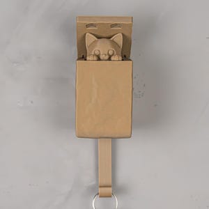 May include: A light brown, rectangular wall hook featuring a sculpted cat head peeking out from the top compartment. The hook has a smooth, matte finish and includes a silver-toned metal ring at the bottom for holding keys or small items.
