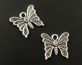 Pode incluir: Dois pingentes de borboleta prateados com detalhes intrincados e um laço para prender em joias ou artesanato.