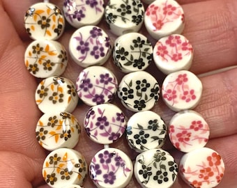 Può includere: Primo piano di una collana di perline di ceramica bianca. Ogni perlina è rotonda e decorata con un motivo floreale in vari colori, tra cui arancione, viola, nero e rosa. Le perline sono infilate con un filo bianco.