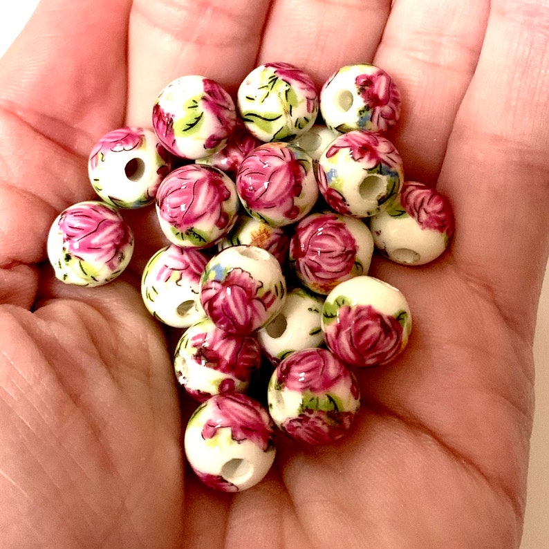 May include: A collection of white ceramic beads with pink and green floral designs. The beads are round and have a hole in the center.