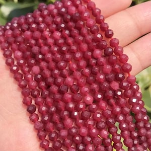 May include: Close-up of strands of faceted, deep red gemstone beads. The beads are small, round, and strung together, showcasing a rich, burgundy color. The beads are held in a hand, suggesting their size and texture. Ideal for jewelry making.