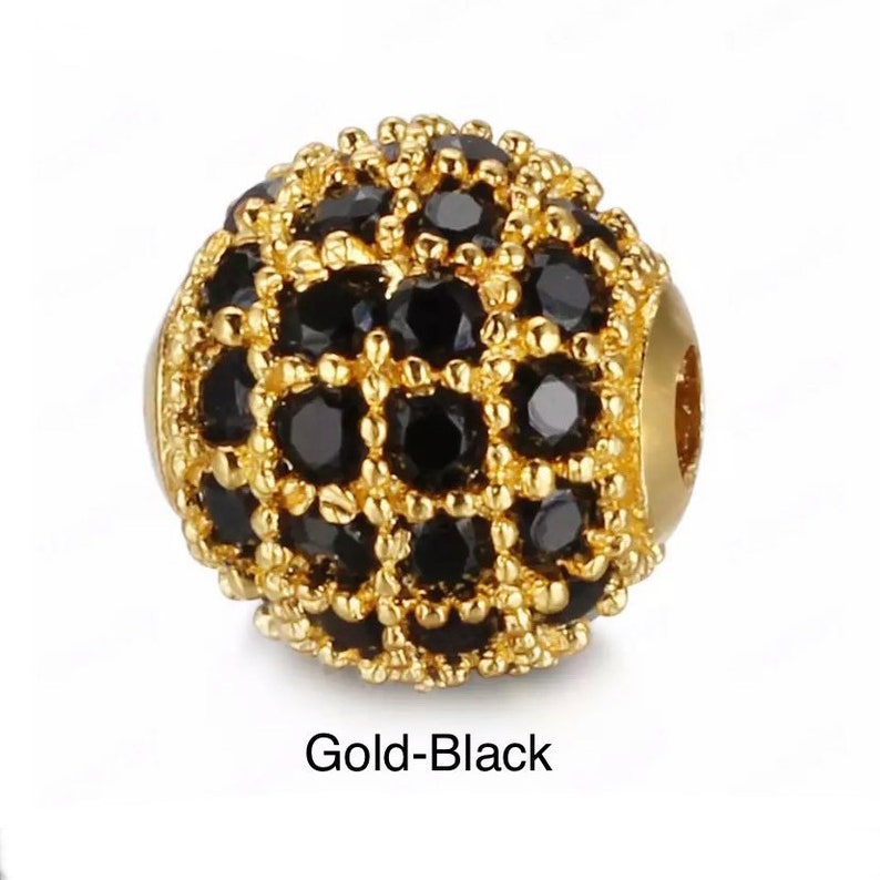 May include: A gold and black round bead with a decorative design of small black stones. The bead has a hole in the center for stringing.