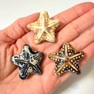 May include: Three ceramic starfish figurines in a hand. The starfish have varying colors and patterns, including beige, brown, and blue. Each starfish has a five-point design with small, white dots.