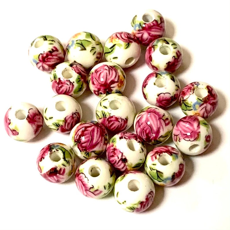 May include: A collection of white ceramic beads with a floral pattern in shades of pink, green, and blue. The beads have a hole in the center for stringing.