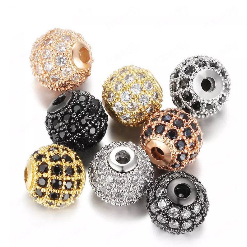 May include: Assortment of round beads with a variety of finishes, including gold, silver, and black. Each bead is covered in small, sparkling rhinestones.