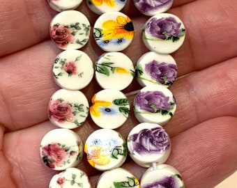 May include: A strand of white ceramic round beads with floral designs. The beads feature various floral patterns, including roses, sunflowers, and other wildflowers. The beads are purple, yellow, and pink.