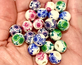 May include: A collection of round, off-white beads with floral designs in shades of pink, blue, and green. The beads are held in a hand, showcasing the variety of patterns and colors. Each bead has a hole for stringing.