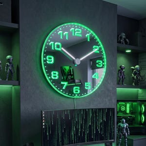 Peut inclure: Une horloge murale circulaire avec une surface miroir réfléchissante et des chiffres et aiguilles LED vert lumineux. L'horloge présente une bordure lumineuse verte de style néon et est fixée sur un mur gris foncé au-dessus d'un bureau informatique.