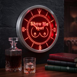May include: A circular wall clock with a silver frame and a glowing red neon face. The clock features the text "Show Me Your" above a line-art graphic of two breasts. The numbers 3, 6, and 9 are displayed in a bold, red, digital-style font.
