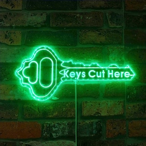 May include: A green neon key-shaped sign with the text "Keys Cut Here" on a brick wall background.