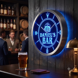 Könnte beinhalten: Eine runde Wanduhr mit einem silberfarbenen Metallrahmen und einem leuchtend blauen LED-Zifferblatt. Die Uhr zeigt den Text DANIEL'S BAR, eine Grafik mit zwei Bierkrügen sowie Ziffern an den Positionen 3, 6 und 9 auf einer dunklen Holzwand.