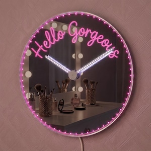 Könnte beinhalten: Eine runde Wanduhr mit verspiegelter Oberfläche und dem pinkfarbenen Neon-Schriftzug Hello Gorgeous. Die Uhr hat einen kreisförmigen Rahmen, der von kleinen pinken LED-Lichtern beleuchtet wird, sowie weiße Zeiger. Der Spiegel reflektiert einen Schminktisch.