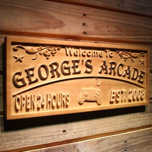 Könnte beinhalten: Ein Holzschild mit dem Text "Willkommen in Georges Arcade Geöffnet 24 Stunden Est. 2008" und einem geschnitzten Bild eines Videospiel-Controllers.
