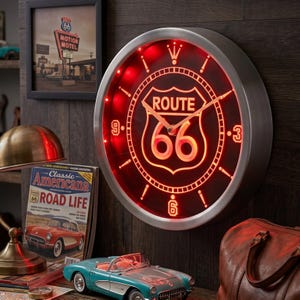 Puede incluir: Un reloj de pared redondo con marco de metal plateado cepillado y esfera negra que presenta un emblema de neón rojo brillante de la Ruta 66. El reloj tiene manecillas de metal finas, marcadores de hora iluminados en rojo y los números 3, 6 y 9.