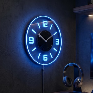 Puede incluir: Un reloj de pared circular con una esfera central negra y manecillas plateadas, rodeado por una cara transparente con números 3, 6, 9 y 12 brillantes en azul neón, y un anillo de luz azul continuo alrededor del borde exterior del marco.