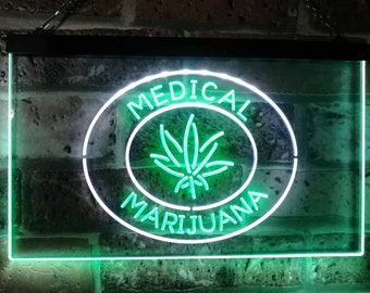 Puede incluir: Un letrero de neón rectangular con un marco negro, que muestra las palabras "MEDICAL MARIJUANA" en blanco y verde. El letrero presenta un emblema de hoja de cannabis verde dentro de un óvalo blanco. El fondo es una pared de ladrillos.