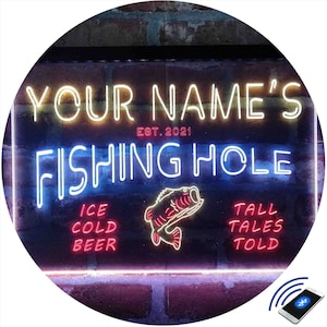 May include: A neon sign with the text "YOUR NAME'S FISHING HOLE EST. 2021" in white and blue neon. The sign also features a red neon fish, and the text "ICE COLD BEER" and "TALL TALES TOLD" in red neon.