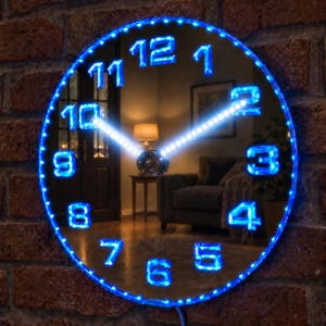 May include: A circular wall clock with a mirrored surface and a blue LED light border. The clock features glowing blue digital-style numerals from 1 to 12 around the perimeter and two bright white LED hands indicating the time on the reflective face.