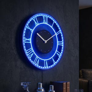 Puede incluir: Un reloj de pared circular con una esfera central negra, manecillas plateadas y un anillo de neón azul brillante. El borde exterior muestra números romanos blancos sobre un fondo oscuro, creando una pantalla iluminada de alto contraste.
