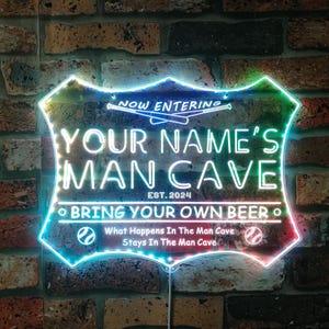 May include: A neon sign with a baseball theme. The sign says "Now Entering Your Name's Man Cave Est. 2024 Bring Your Own Beer" with baseball bats and baseballs. The sign is lit up with multi-colored lights.