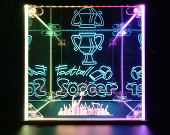 Puede incluir: Una caja de acrílico transparente con un brillo verde neón y morado. La caja presenta un trofeo, las palabras "Football" y "Soccer", y un grupo de figuras en miniatura levantando las manos en señal de celebración.