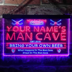 May include: Neon sign with the text "NOW ENTERING YOUR NAME'S MAN CAVE EST. 2019 • BRING YOUR OWN BEER • What Happens In The Man Cave Stays In The Man Cave" in pink and blue neon lettering. The sign has two crossed hockey sticks on either side of the text.