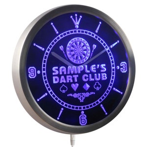 May include: A silver-colored wall clock with a black face and blue LED lights. The clock features a dartboard, darts, and the text "SAMPLE'S DART CLUB".