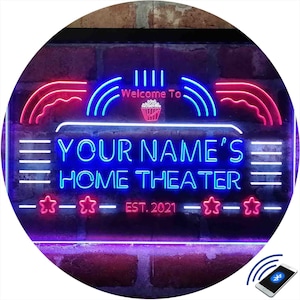 May include: A blue and pink neon sign with the text "Welcome To YOUR NAME'S HOME THEATER EST. 2021" with a popcorn bucket icon. The sign is on a brick wall and has a Bluetooth icon in the bottom right corner.