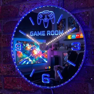May include: A circular wall clock with a blue LED-lit frame and a mirrored surface. The clock face features a glowing blue game controller icon at the top, the text "GAME ROOM" in the centre, and blue illuminated numbers at the 3, 6, and 9 positions.