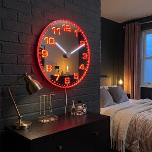 Puede incluir: Un reloj de pared circular con cara de espejo y un borde de luz LED rojo brillante. El reloj presenta grandes números rojos brillantes de estilo digital del 1 al 12 y dos manecillas blancas iluminadas. Un cable cuelga de la parte inferior.