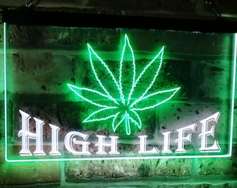 Puede incluir: Un letrero de neón rectangular con un diseño de hoja de cannabis verde y las palabras "HIGH LIFE" en blanco. El letrero está montado en una pared de ladrillo, y la luz de neón emite un brillo verde brillante.