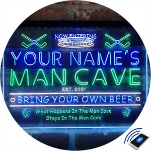 May include: A blue and green neon sign with the text "NOW ENTERING YOUR NAME'S MAN CAVE EST. 2021 • BRING YOUR OWN BEER • What Happens In The Man Cave Stays In The Man Cave". The sign is illuminated and has a remote control.