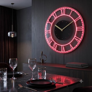 Può includere: Un orologio da parete circolare con quadrante centrale nero, lancette dorate e una cornice luminosa al neon rosso. La cornice presenta grandi numeri romani in luce neon rossa, creando un bordo circolare luminoso sulla parete scura.