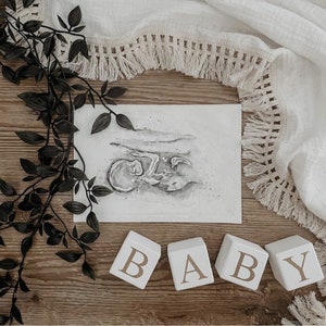 May include: Four white wooden blocks spelling out the word "BABY" in gold lettering. The blocks are on a wooden surface with a black and white ultrasound image and a white fringed fabric in the background.