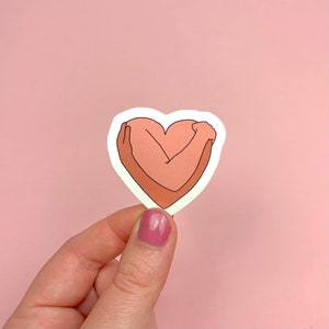 May include: A sticker featuring a heart shape with arms embracing it. The arms and heart are in shades of pink and peach, outlined in black. The sticker is held against a pink background.