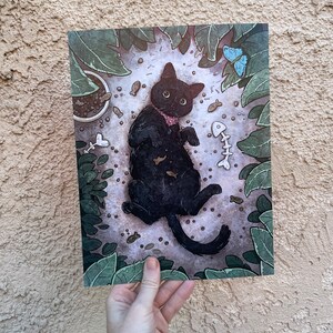 Puede incluir: Un gato negro con un collar rosa yace de espaldas rodeado de hojas verdes y huesos de pescado y otros juguetes para gatos esparcidos.