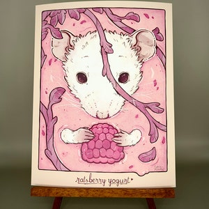May include: An illustration of a white rat holding a large raspberry in a pink liquid, with the text "ratsberry yogurt" at the bottom. The artwork features purple branches and leaves, creating a whimsical scene.